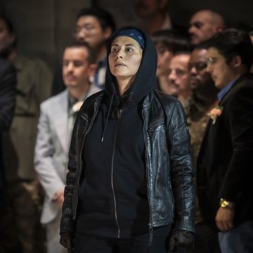 Tancredi aus Rossinis Tancredi steht im Mittelpunkt einer Gruppe von Menschen. Die Figur trägt dunkle Kleidung mit Kapuzenjacke, Lederjacke, Handschuhen und einem blauen Kopftuch. Mit ernstem, konzentriertem Blick schaut Tancredi nach oben, während die Personen im Hintergrund unscharf bleiben. Das Bühnenlicht lenkt die Aufmerksamkeit auf die isoliert wirkende Figur und unterstreicht ihre innere Anspannung. Die Szene vermittelt Entschlossenheit, Einsamkeit und die ständige Gefahr eines Lebens im Verborgenen innerhalb der von rivalisierenden Clans geprägten Welt dieser Inszenierung.