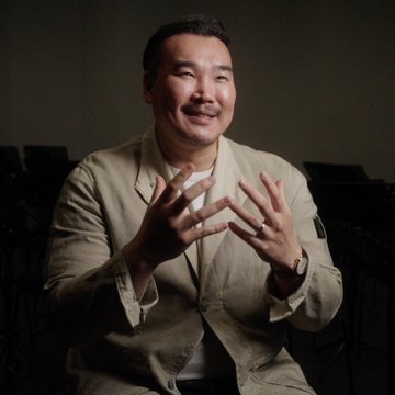 Porträt von Young Woo Kim während eines Interviews: Der Sänger sitzt vor dunklem Hintergrund, lächelt und gestikuliert mit beiden Händen, als würde er lebhaft über seine Rolle sprechen.