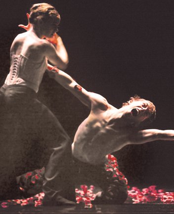 Zwei Tänzer befinden sich in einer intensiv inszenierten Bühnenszene mit dunklem Hintergrund und stark fokussiertem, warmem Licht, das ihre Körper plastisch hervorhebt.
Im Vordergrund kniet ein oberkörperfreier Tänzer auf dem Boden, sein Oberkörper ist nach hinten gebogen, die Arme weit ausgebreitet. Sein Kopf ist weit zurückgeworfen, der Mund geöffnet – der Ausdruck wirkt wie ein Schrei oder ein Moment äußerster emotionaler oder körperlicher Anspannung. Links neben ihm steht eine Tänzerin, die ein weißes, eng geschnürtes Korsett und dunkle Hosen trägt. Sie beugt sich nach vorne und greift den knienden Tänzer am Arm, wodurch eine Verbindung zwischen beiden entsteht, die zugleich unterstützend und kontrollierend wirkt. Auf dem Boden liegen zahlreiche rote Blütenblätter, die einen starken farblichen Kontrast zur ansonsten reduzierten, dunklen Szene bilden. Die gesamte Komposition wirkt hochgradig expressiv und körperbetont, fast skulptural, und vermittelt eine Szene von Spannung, Hingabe oder Konflikt zwischen den beiden Tänzern.