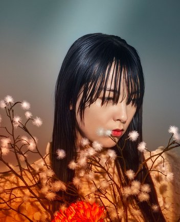 Nahaufnahme einer jungen asiatischen Frau mit cineastischer Bildwirkung und warmer Lichtstimmung: Eine junge Frau mit sehr langem, glattem, schwarzem Haar und dichtem Pony ist im Halbprofil dargestellt und blickt mit gesenktem, nachdenklichem Ausdruck zur rechten Seite. Ihr Gesicht ist weich und warm ausgeleuchtet, die Lippen sind rot bemalt und betont. Sie trägt ein helles, zart strukturiertes Gewand. Im Vordergrund rahmen feine, verzweigte Äste mit kleinen, weißen, flauschigen Blüten ihr Gesicht ein. Unten im Bild setzt eine leuchtend rote Blüte einen starken Farbkontrast. Der Hintergrund ist weich verschwommen in kühlen Grau- und Blautönen, wodurch die Figur und die warmen Farben im Vordergrund hervorgehoben werden. Die Gesamtstimmung wirkt still, melancholisch und poetisch. Die Szene evoziert die Figur der Cio-Cio-San aus Giacomo Puccinis Oper Madama Butterfly.