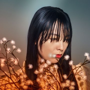 Close-up of a young Asian woman with a cinematic look and warm lighting: A young woman with very long, straight black hair and full bangs is shown in half profile, gazing pensively to the right with a lowered expression. Her face is softly and warmly illuminated, with red-painted, accentuated lips. She wears a light, delicately textured garment. In the foreground, fine, branching twigs with small, white, fluffy blossoms frame her face. At the bottom of the image, a vivid red flower creates a strong color contrast. The background is softly blurred in cool shades of grey and blue, highlighting the figure and the warm tones in the foreground. The overall mood is quiet, melancholic, and poetic. The scene evokes the character of Cio-Cio-San from Madama Butterfly by Giacomo Puccini.