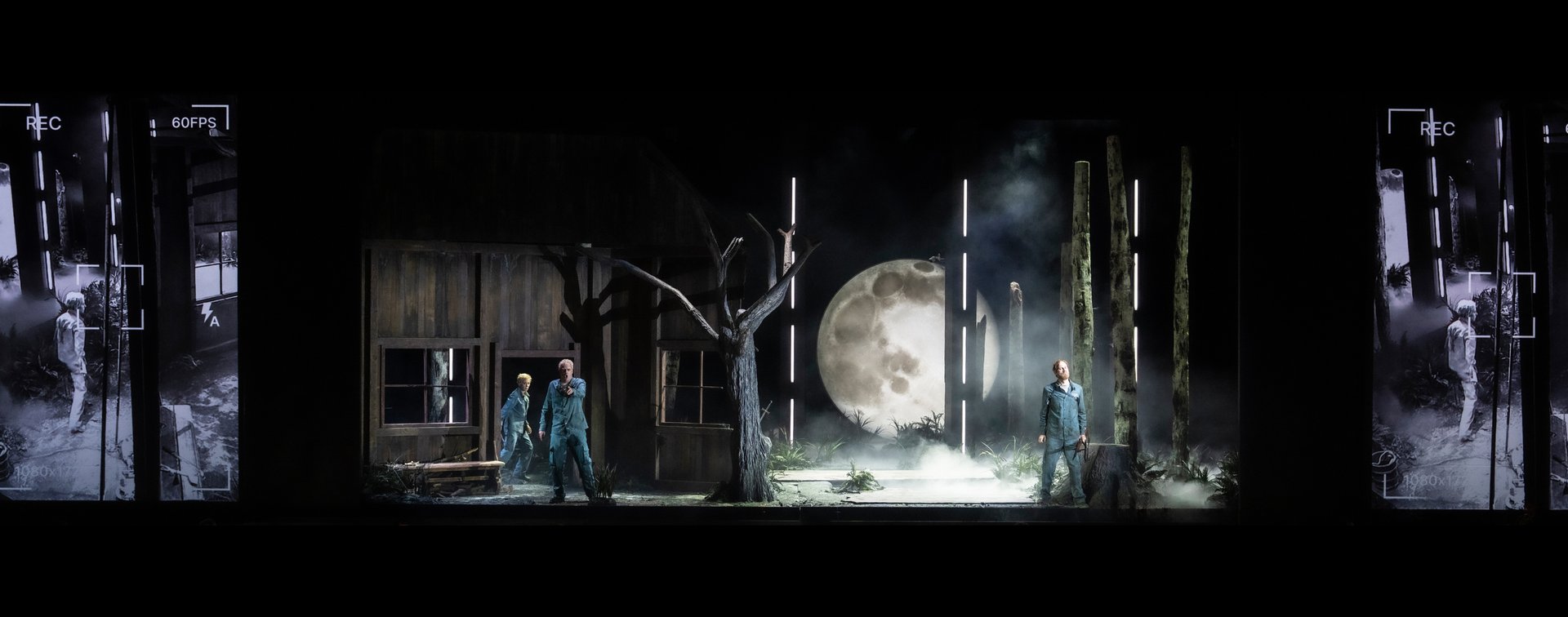 Auf einer düsteren Bühnenlandschaft stehen drei Figuren – Sieglinde, Siegmund und Hunding – aus Wagners Die Walküre. Links ein schlichtes Holzhaus, davor stehen Sieglinde und Siegmund in grüner Kleidung. Rechts steht Hunding etwas abseits zwischen stilisierten, senkrechten Lichtstäben, die einen Wald andeuten. Im Hintergrund dominiert ein großer, heller Vollmond, der die Szene in kaltes Licht taucht. Seitlich sind schwarz-weiße Videoprojektionen mit Überwachungsästhetik zu sehen.
