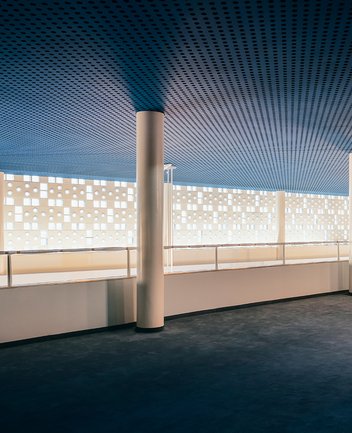 Zweites Geschoss des großzügigen Foyerbereichs der Oper Köln am Offenbachplatz mit hoher Decke und weiter Fensterfront. Die Wand ist von vielen kleinen runden und quadratischen Öffnungen durchzogen, durch die Tageslicht einfällt. Schlanke, helle Säulen tragen die Decke, die in einem kühlen Blauton gestaltet ist und einen Kontrast zur warmen Innenarchitektur bildet.