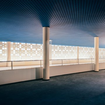 Zweites Geschoss des großzügigen Foyerbereichs der Oper Köln am Offenbachplatz mit hoher Decke und weiter Fensterfront. Die Wand ist von vielen kleinen runden und quadratischen Öffnungen durchzogen, durch die Tageslicht einfällt. Schlanke, helle Säulen tragen die Decke, die in einem kühlen Blauton gestaltet ist und einen Kontrast zur warmen Innenarchitektur bildet.