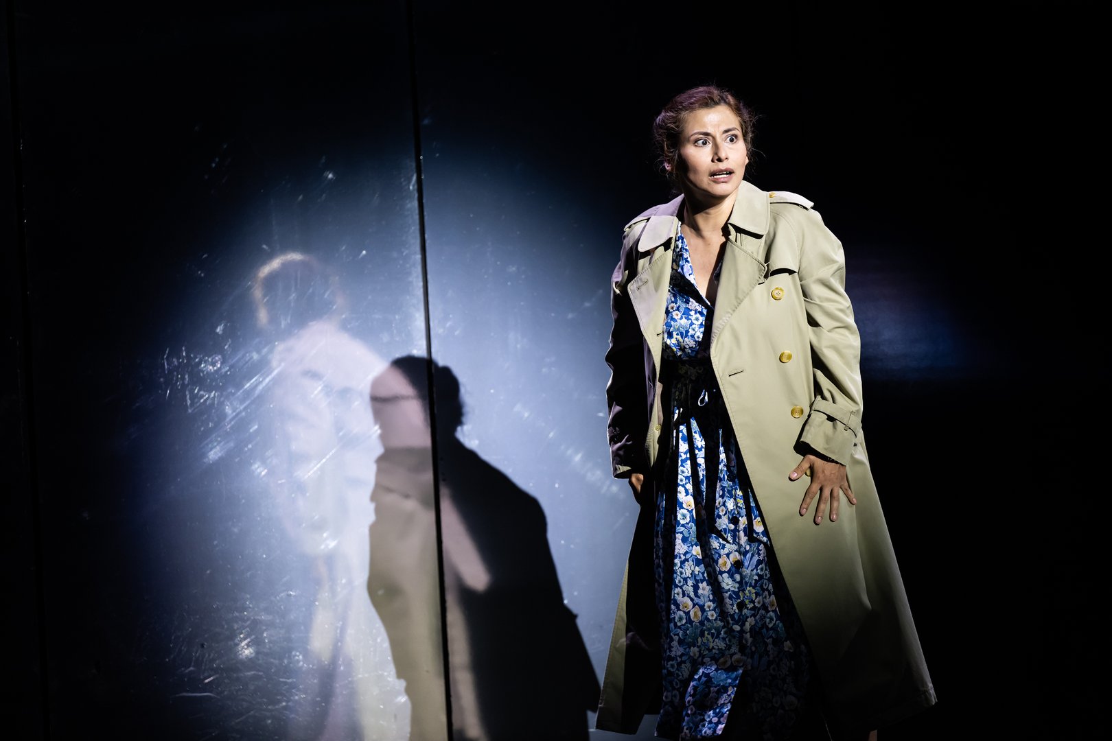 Die Woman aus der Oper Picture a day like this steht allein in einer dunkel gehaltenen Bühnenszene. Sie trägt einen hellen Trenchcoat über einem blau gemusterten Kleid und blickt mit weit geöffneten Augen angespannt zur Seite. Hinter ihr spiegeln sich ihre Silhouette und Teile ihres Gesichts in einer glänzenden Wandfläche. Das starke Seitenlicht hebt die Figur deutlich aus dem schwarzen Hintergrund hervor und erzeugt eine intensive, dramatische Stimmung.
