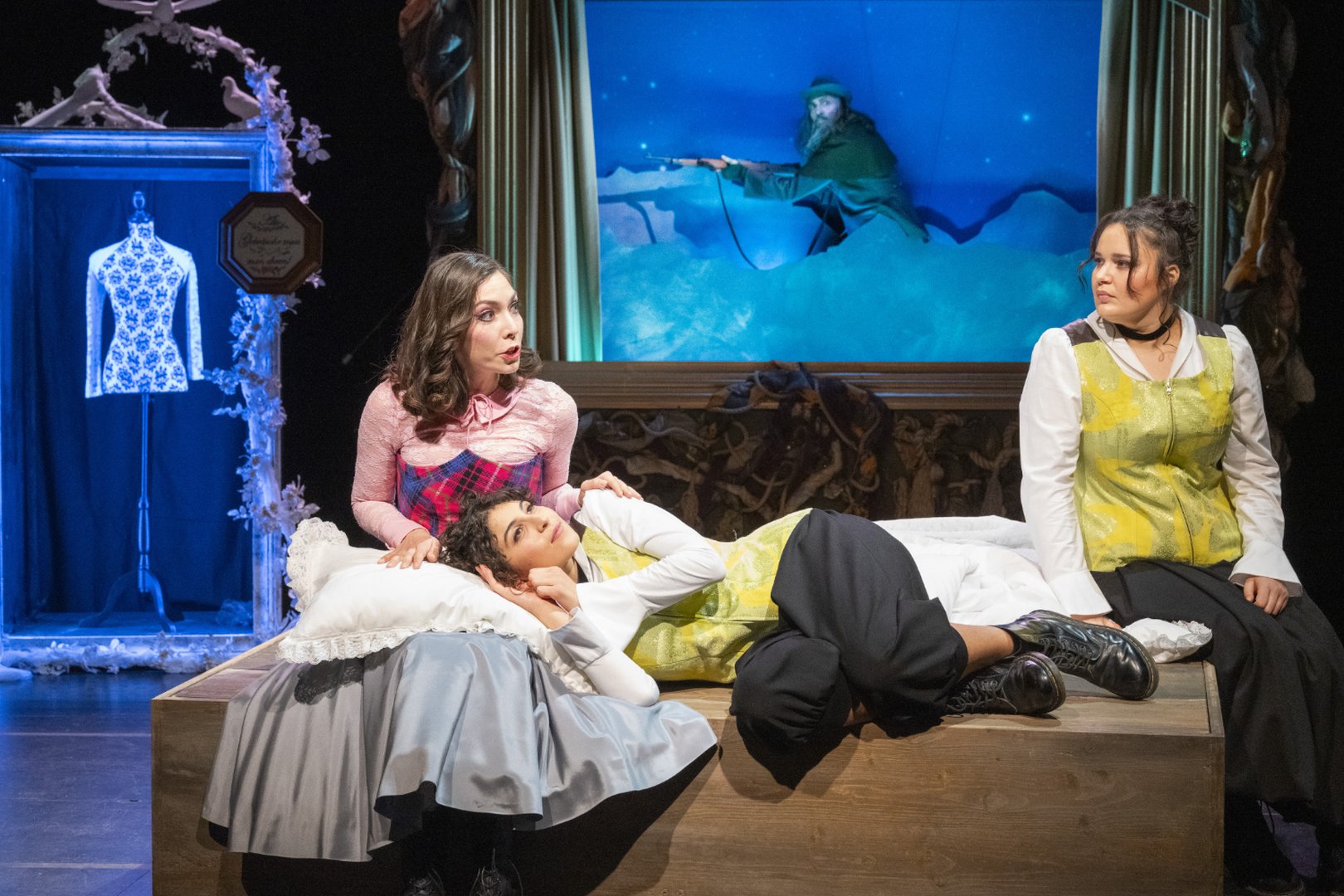 Vordergrund liegt die Darstellerin der Agathe mittig auf einer riesigen Holzbox, den Kopf auf dem Schoss von Ännchen auf einem Kissen abgestützt, in einem gelb gemusterten Oberteil, weißer Bluse und schwarzen Hosen. Sie wirkt nachdenklich und ruhig. Diese Darstellerin nutzt Deutsche Gebärdensprache (DGS) und macht die Figur Agathe zweisprachig erfahrbar.
Links neben ihr sitzt Ännchen in einem auffälligen Kostüm aus rosa Oberteil und gemustertem Kleid. Sie beugt sich leicht zu Agathe hinunter und tätschelt sie, während sie zur rechten Person, ebenso Agathe, blickt. Sie spricht zu beiden. Rechts sitzt die zweite Darstellerin der Agathe, ebenfalls in gelb gemusterter Weste und weißer Bluse, und blickt aufmerksam zur Mitte der Szene.
Im Hintergrund ist ein großes, rahmenartiges Bühnenbild zu sehen, das eine winterliche Szene zeigt: Erbförster Kuno mit Gewehr bewegt sich durch eine verschneite Landschaft unter einem blauen, nächtlichen Himmel. Links steht ein dekorativer Rahmen mit einem ausgestellten Kleid. Die Szene stammt aus der Oper Freikugeln.