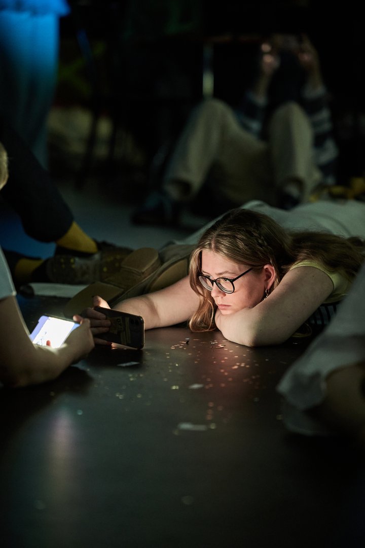 Szene aus When I find myself in Times of Trouble des Spielclubs Opera, Song and Scene. Eine Person mit Brille liegt auf dem Boden der Bühne und stützt ihren Kopf auf die verschränkten Arme. Vor ihr liegt ein Smartphone, dessen Bildschirm ihr Gesicht schwach beleuchtet. Ihr Blick ist konzentriert auf das Gerät gerichtet, während die Umgebung im Halbdunkel verschwimmt. Im Vordergrund ist die Hand einer weiteren Person mit einem leuchtenden Smartphone zu sehen. Im Hintergrund sitzen und liegen weitere Mitwirkende verstreut im Raum. Kleine helle Partikel auf dem Boden reflektieren das Bühnenlicht und verstärken die ruhige, nachdenkliche Atmosphäre der Szene.
