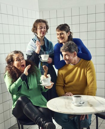 In einem hellen, schlicht gefliesten Raum – der an ein Café oder eine Küche erinnert – sitzt eine Gruppe von vier Erwachsenen eng beieinander an einem kleinen runden Tisch. Die Szene wirkt bewusst inszeniert und leicht surreal.
Im Vordergrund links sitzt eine Frau in einem grünen Oberteil und schwarzen Lederhosen auf einem Stuhl. Sie hält eine weiße Tasse mit Untertasse in der Hand, aus der Sahne quillt – und aus dieser Sahne ragt überraschend eine Schlange empor. Ihr Gesichtsausdruck ist stark überzeichnet: Mit weit aufgerissenen Augen und offenem Mund weicht sie erschrocken zurück, als würde sie die Situation gerade erst bemerken.

Direkt hinter ihr steht eine weitere Frau mit kurzem Haar und einem Tuch um die Schultern. Sie hält ebenfalls eine Tasse sowie einen Löffel in der Hand und wirkt im Gegensatz dazu ruhig und beinahe ungerührt, als beobachte sie das Geschehen mit nüchterner Neugier.

Rechts im Bild sitzt ein Mann in einem senffarbenen Pullover an dem kleinen Tisch. Vor ihm steht eine weitere Tasse Kaffee. Er dreht sich leicht zur Szene hin und schaut mit einem amüsierten, fast schelmischen Lächeln auf die erschrockene Frau.

Hinter ihm lehnt sich eine dritte Frau in einem blauen Oberteil über seine Schulter. Sie lächelt warm und scheint die Situation ebenfalls eher unterhaltsam als beängstigend zu finden.

Die Kontraste zwischen den Reaktionen – Schreck, Gelassenheit und Belustigung – sowie das absurde Detail der Schlange in der Kaffeetasse verleihen dem Bild eine humorvolle, leicht theatralische und inszenierte Atmosphäre.