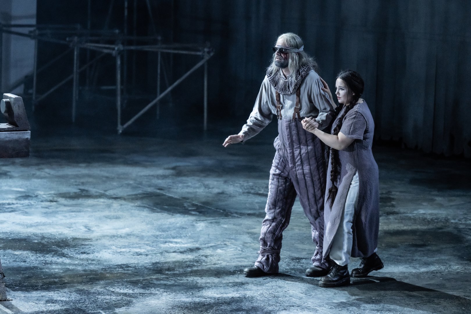 Zu sehen sind Timur und Liù in einer ruhigen Szene aus „Turandot“. Liù führt den blinden Timur vorsichtig über die Bühne und hält seine Hand, während er tastend den Weg sucht. Beide tragen schlichte, grauviolette Kostüme, die die düstere und melancholische Stimmung der Szene unterstreichen. Das kalte Bühnenlicht und die weite, fast leere Bühne lenken den Fokus auf die enge Verbindung der beiden Figuren.