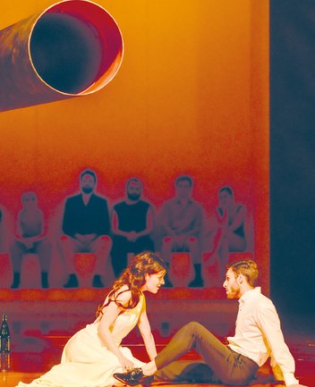 Poppea und Nero in einer Szene aus Die Krönung der Poppea von Claudio Monteverdi: Im Vordergrund kniet Poppea in einem hellen, fließenden Kleid auf dem Bühnenboden und beugt sich intensiv zu Nero hinüber, der ihr gegenüber sitzt, sich nach hinten abstützt und sie aufmerksam ansieht. Die Szene ist in warmes, orangefarbenes Licht getaucht und wirkt sinnlich und spannungsgeladen. Im Hintergrund, leicht erhöht und unscharf dargestellt, sitzt eine Reihe von Figuren nebeneinander und beobachtet das Geschehen. Über ihnen ragt ein großes, zylinderförmiges Objekt in den Raum hinein, das die reduzierte, moderne Bühnenästhetik unterstreicht.