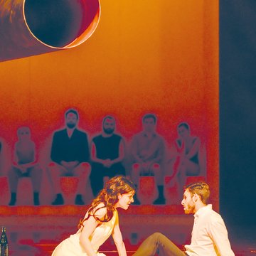 Poppea and Nero in a scene from The Coronation of Poppea by Claudio Monteverdi: In the foreground, Poppea kneels on the stage floor in a light, flowing dress, leaning intently toward Nero, who sits opposite her, supporting himself with his arms behind him and looking at her attentively. The scene is bathed in warm, orange light, creating a sensual and tension-filled atmosphere. In the background, slightly elevated and out of focus, a row of figures sits side by side, observing the action. Above them, a large cylindrical object extends into the space, emphasizing the minimalist, modern stage design.