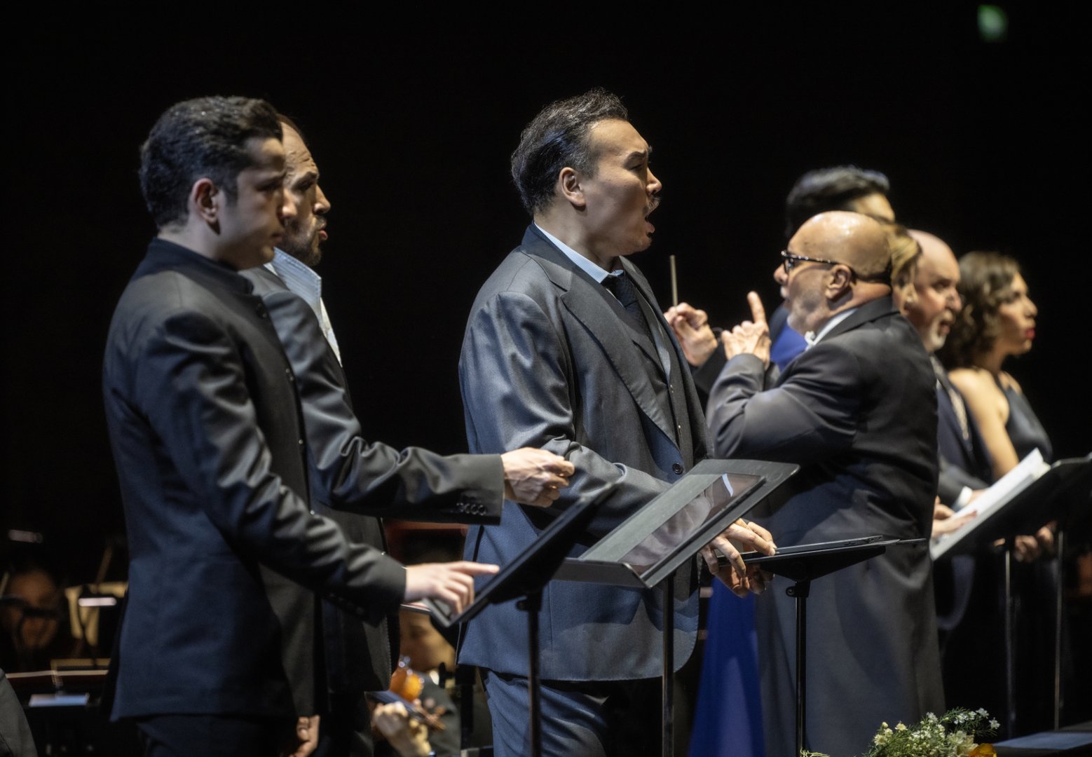 Auf der Bühne stehen Ferhat Baday (Jago), Adrian Sâmpetrean (Don Ruy Gómez de Silva), Young Woo Kim (Ernani), Marta Torbidoni (Elvira), Insik Choi (Don Carlos), John Heuzenroeder (Don Riccardo) und Alina König Rannenberg (Giovanna) nebeneinander an Notenpulten und singen mit ausdrucksstarker Gestik. Im Vordergrund rechts dirigiert Giuliano Carella (musikalische Leitung) mit erhobenem Taktstock und gibt präzise Einsätze, während sich die Sänger*innen auf ihn konzentrieren.