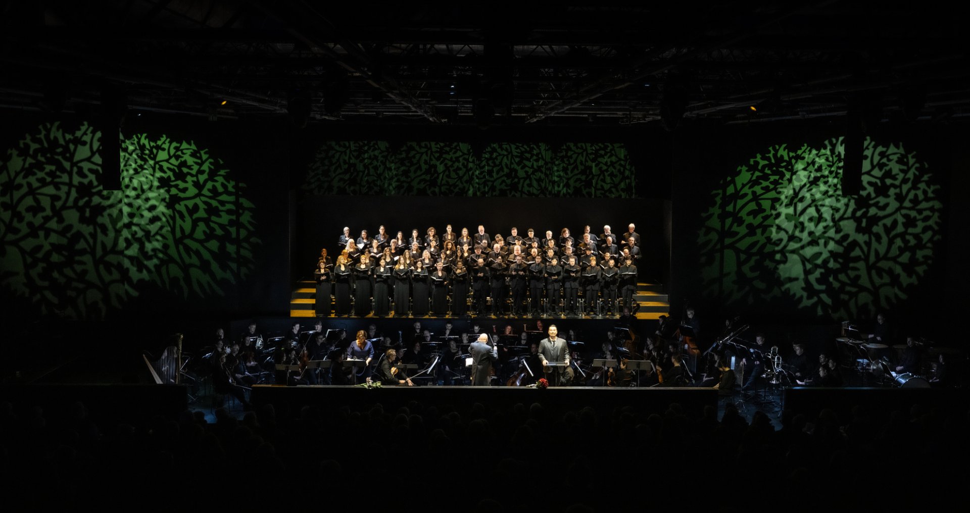 Eine weite Bühnenansicht zeigt das Gürzenich Orchester im Orchestergraben und darüber den Chor der Oper Köln auf gestaffelten Podesten. In der Mitte dirigiert Giuliano Carella (musikalische Leitung) das Ensemble. Im Vordergrund stehen Marta Torbidoni (Elvira) und Young Woo Kim (Ernani) an Notenpulten und singen, während das Bühnenbild mit grünen, baumartigen Lichtprojektionen die Szene rahmt.