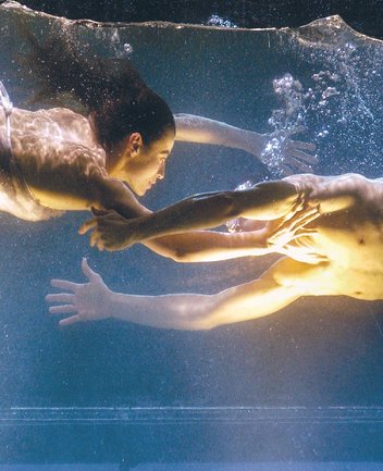 Unterwasserszene aus Dido und Aeneas: Zwei Darstellende schweben einander gegenüber in einem Wasserbecken, ihre Körper sanft im Raum gehalten, die Arme einander entgegenstreckend, während Luftblasen zwischen ihnen aufsteigen und warmes, goldenes Licht die Szene durchzieht.