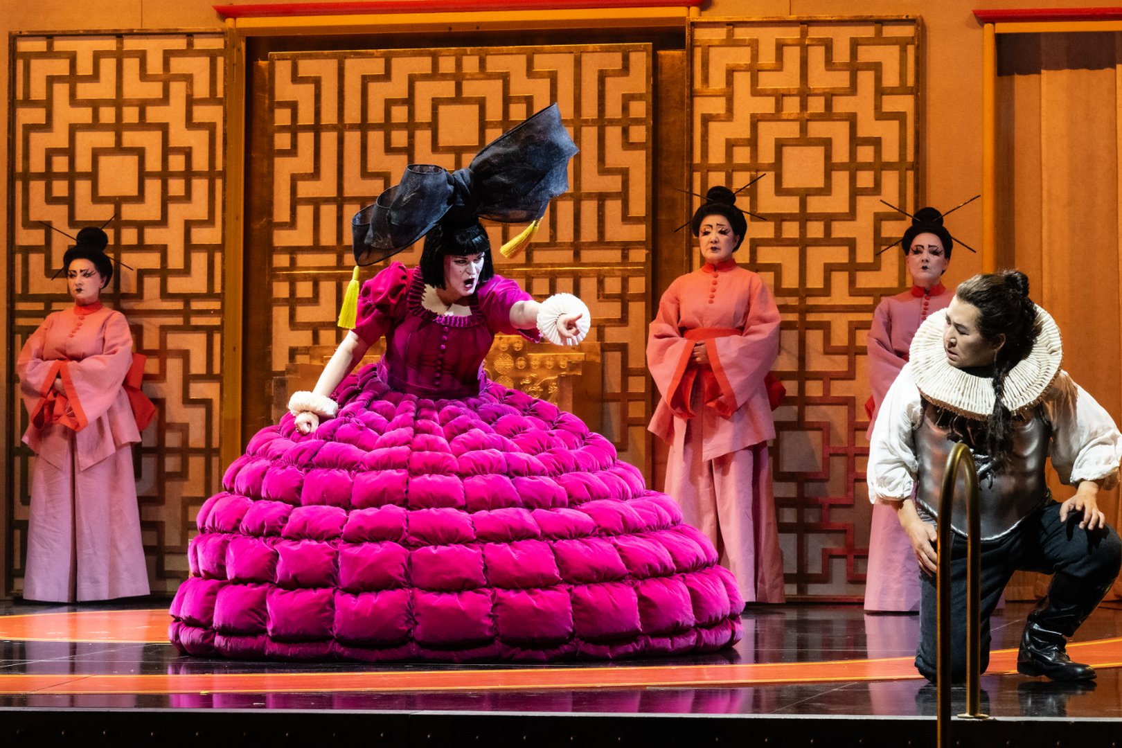 Zu sehen sind Turandot und Calaf in einer intensiven Szene aus „Turandot“. Turandot sitzt in einem auffälligen, voluminösen pinkfarbenen Kleid mit übergroßer schwarzer Schleife vor einem ornamentalen Hintergrund und zeigt energisch auf Calaf. Dieser kniet ihr gegenüber in dunkler Kleidung mit weißem Halskragen und blickt herausfordernd zu ihr auf. Drei Dienerinnen in rosafarbenen Gewändern rahmen die Szene symmetrisch ein. Die kräftigen Farben und die stilisierte Ausstattung verleihen der Inszenierung eine zugleich märchenhafte und theatralische Wirkung.