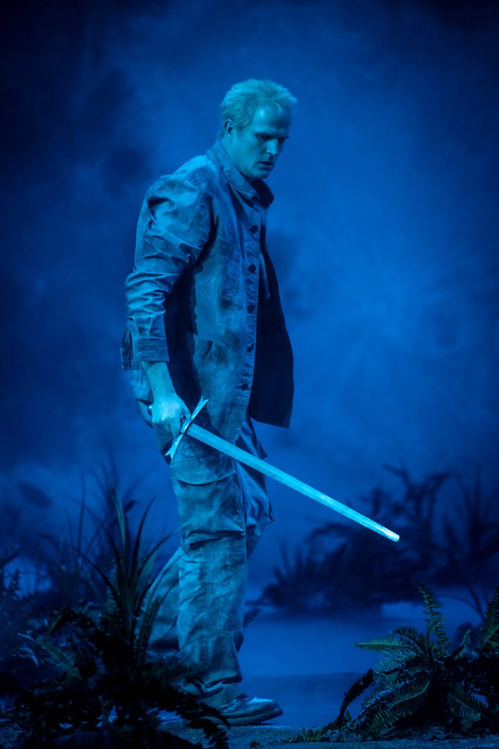 In bläuliches Licht und Nebel getaucht steht Siegmund mit ernstem, angespanntem Blick auf einer kargen Bühne und hält ein Schwert schräg nach unten gerichtet in der Hand. Die Szene wirkt kühl, still und bedrohlich und zeigt eine zentrale Figur aus Die Walküre.