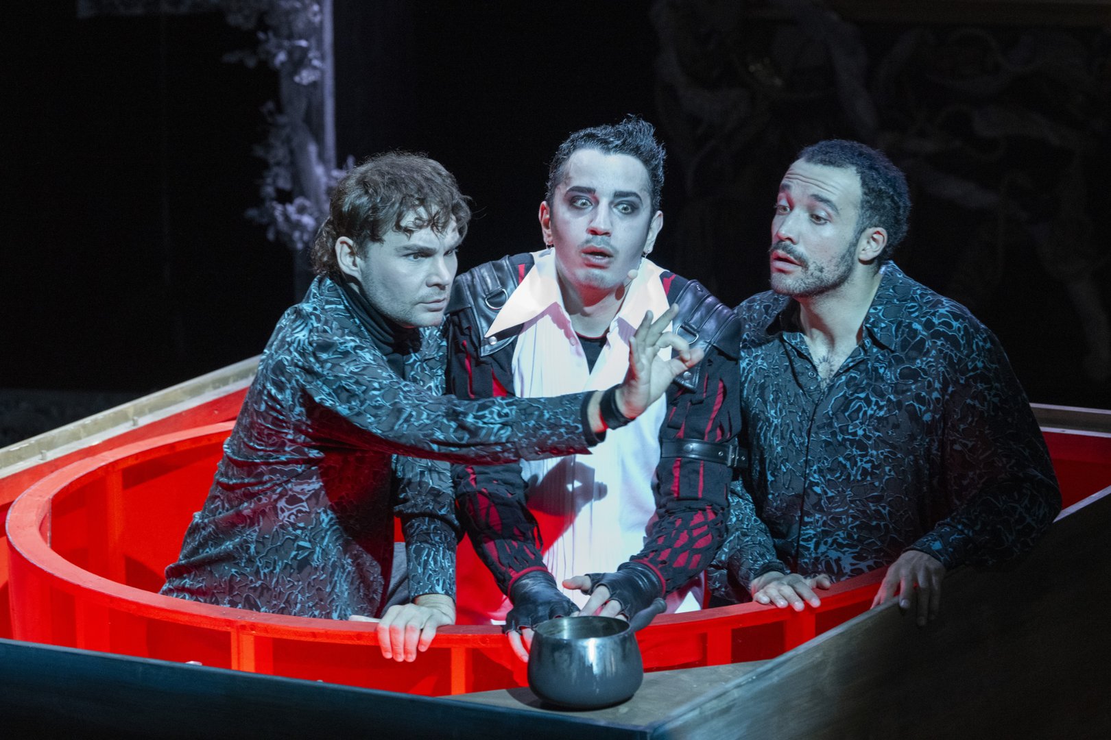Drei Darsteller stehen dicht beieinander in einer intensiven, düster ausgeleuchteten Szene auf der Bühne. Sie befinden sich in einer runden, rot leuchtenden Konstruktion, die wie ein Kessel oder eine magische Vorrichtung wirkt. In der Mitte steht Kaspar in einem auffälligen Kostüm mit schwarzer Lederjacke, roten Elementen und weißem Hemd. Sein Gesicht ist stark ausgeleuchtet, die Augen weit geöffnet, wodurch er angespannt und fast entrückt wirkt. Vor ihm steht ein kleiner metallener Topf.
Links neben ihm steht der Schauspieler der Figur Max, der Deutsche Gebärdensprache (DGS) nutzt. Er beugt sich leicht nach vorne und führt eine präzise Handbewegung aus, die Teil der Gebärdensprache ist, während er konzentriert auf das Geschehen blickt. Rechts steht der Sänger der Figur Max, ebenfalls aufmerksam und angespannt, und beobachtet Kaspar und die Handlung im Zentrum.
Die Szene ist von starkem Kontrast geprägt: kaltes blaues Licht auf den Figuren und ein intensives rotes Leuchten aus der Konstruktion, was eine bedrohliche, magische Atmosphäre erzeugt. Die Darstellung stammt aus der Oper Freikugeln und zeigt eine zentrale, unheimliche Interaktion zwischen Kaspar und Max.