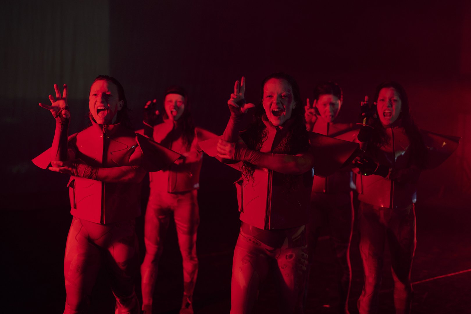 Fünf Tänzer*innen in futuristisch wirkenden Kostümen bewegen sich in einer Reihe auf einer intensiv rot beleuchteten Bühne nach vorne. Im Vordergrund formen sie mit ihren Händen markante, spannungsgeladene Gesten: Finger sind gespreizt, gekrümmt oder gezielt ausgestreckt, teilweise wie Krallen oder präzise Zeichen, die nach außen gerichtet sind. Die Hände wirken kontrolliert und ausdrucksstark, als würden sie etwas greifen, zeigen oder projizieren. Die Münder sind geöffnet, als würden sie rufen. Insgesamt entsteht eine kraftvolle, energetische und leicht aggressive Wirkung mit starkem Fokus auf die choreografierte Handarbeit.