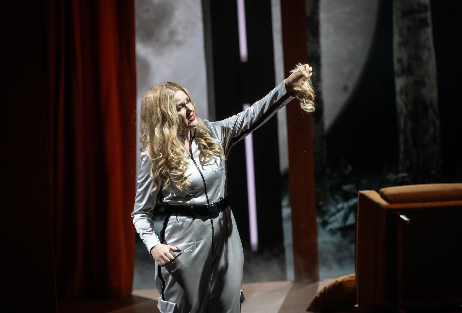 Auf einer dramatisch ausgeleuchteten Bühne hält Brünnhilde eine abgeschnittene Haarsträhne in die Höhe, während sie in der anderen Hand eine Schere trägt. Ihr Ausdruck ist intensiv und entschlossen. Hinter ihr sind rote Vorhänge, Nebel und angedeutete Bühnenelemente zu sehen, die die spannungsgeladene Szene aus Die Walküre unterstreichen.