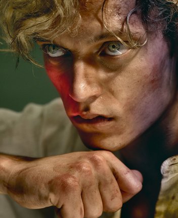 Nahaufnahme eines jungen Mannes mit cineastischer Bildwirkung und warmer Lichtstimmung: Mit blondem, zerzaustem Haar und schmutzverschmierter Haut blickt er mit grün-blauen Augen intensiv nach vorn. Sein Ausdruck ist angespannt und innerlich aufgewühlt, die rechte Hand zur Faust geballt und nahe am Kinn. Er trägt ein weißes Langarmshirt. Die Szene evoziert die Figur des Kain aus Alessandro Scarlattis Oper Kain und Abel.