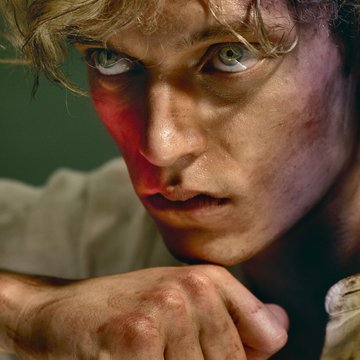 Close-up of a young man with a cinematic look and warm lighting: With tousled blond hair and dirt-smudged skin, he gazes intensely ahead with green-blue eyes. His expression is tense and emotionally unsettled, his right hand clenched into a fist and held close to his chin. He wears a white long-sleeved shirt. The scene evokes the figure of Cain from Alessandro Scarlatti’s opera Cain and Abel.