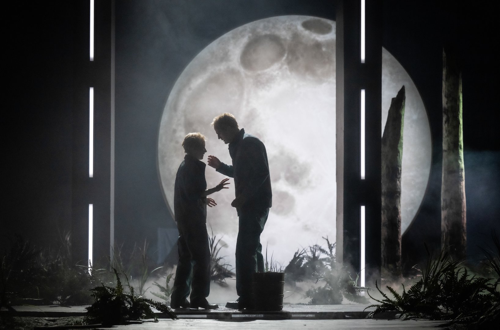 Siegmund und Sieglinde stehen sich als Silhouetten gegenüber, eingerahmt von schmalen, vertikalen Lichtstreifen. Hinter ihnen dominiert ein übergroßer, leuchtender Vollmond die Szene. Nebel liegt über dem Boden, zwischen Farnen und Baumstämmen, und verstärkt die geheimnisvolle, spannungsgeladene Atmosphäre ihres ersten Aufeinandertreffens in Wagners DIE WALKÜRE.