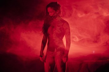 Eine Tänzerin steht allein auf einer intensiv rot ausgeleuchteten Bühne, umgeben von dichtem, wirbelndem Nebel. Ihr Körper ist leicht zur Seite gedreht, der Blick nach unten gerichtet. Die Arme hängen locker herab, die Hände entspannt und geöffnet, mit leicht gekrümmten Fingern, die kaum Spannung zeigen – im Gegensatz zu den zuvor kraftvollen Gesten wirkt diese Haltung ruhig und zurückgenommen. Das lange, dunkle Haar fällt über eine Schulter und bewegt sich leicht im Nebel. Die Szene wirkt still, fast entrückt, mit einer geheimnisvollen, schwebenden Atmosphäre.