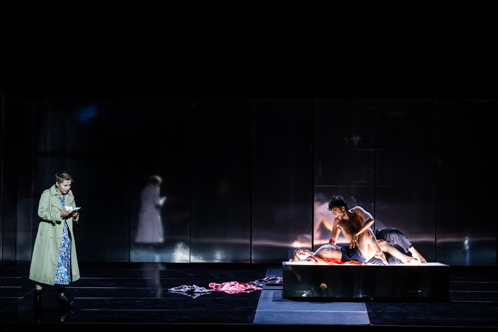 In einer dunkel gehaltenen Bühnenszene steht links die Woman in einem langen beigen Mantel und liest nachdenklich in einem kleinen Buch oder Heft. Rechts liegt Lover 1 / Composer auf einer erhöhten Plattform, während Lover 2 / Composer’s Assistant sich eng über sie beugt. Die beiden sind intensiv beleuchtet, wodurch ihre Körper und Gesten stark hervortreten. Im Hintergrund spiegeln dunkle Paneele die Figuren schemenhaft wider und verstärken die melancholische, intime Atmosphäre der Szene aus der Oper Picture a day like this.
