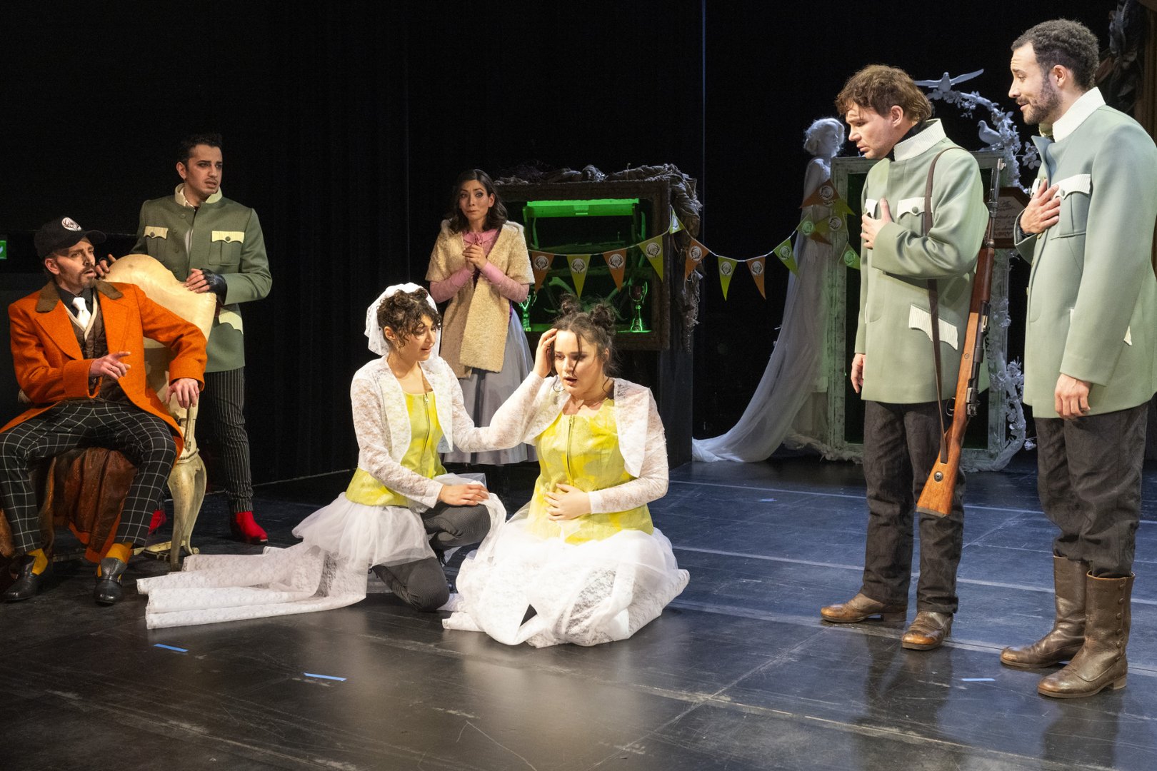 Auf einer Theaterbühne ist eine größere Gruppe von Figuren in einer dramatischen Szene versammelt. Im Zentrum kniet die Sängerin der Agathe in einem weißen Kleid mit gelb gemusterter Weste und wirkt erschüttert, eine Hand an den Kopf gelegt. Neben ihr kniet die Schauspielerin der Agathe, die sie unterstützt und tröstend berührt; sie nutzt Deutsche Gebärdensprache (DGS) und macht die Figur zweisprachig erfahrbar.
Links sitzt Ottokar in einem auffälligen orangefarbenen Mantel auf einem Stuhl und beobachtet die Szene. Hinter ihm steht Kaspar in dunkler Kleidung und wirkt angespannt. In der Mitte im Hintergrund steht Ännchen, die die Hände vor der Brust hält und besorgt auf Agathe blickt.
Rechts stehen zwei Darsteller der Figur Max nebeneinander: der Sänger und der Schauspieler. Der Schauspieler nutzt DGS und trägt ein Gewehr über der Schulter. Beide wirken betroffen und blicken in Richtung Agathe.
Das Bühnenbild zeigt im Hintergrund dekorative Elemente wie einen Rahmen mit einem weißen Kleid und eine grün beleuchtete Nische. Die Szene stammt aus der Oper Freikugeln und zeigt eine konflikthafte, emotional aufgeladene Situation zwischen den zentralen Figuren.