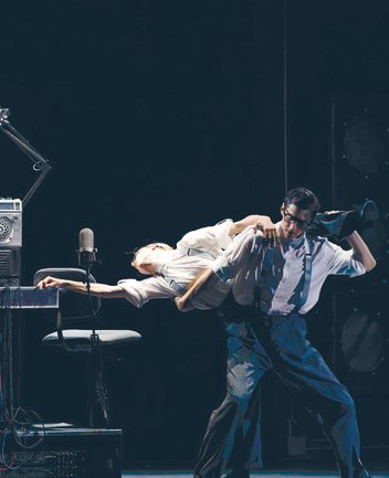 Mehrere Tänzer in einer stilisierten Bühnenszene aus der Choreografie Notte Morricone: Im Zentrum trägt ein männlicher Tänzer mit Brille, Hemd und Hosenträgern eine Tänzerin quer über seinen Schultern, während sie ihren Oberkörper nach hinten streckt und einen Arm ausstreckt. Die Szene ist von Requisiten wie einem Mikrofon, einem Stuhl und technischem Equipment geprägt, was eine Atmosphäre zwischen Bühne und Studio erzeugt.