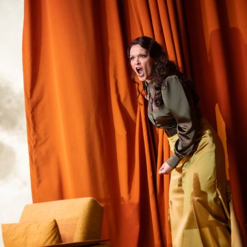 Auf einer modern gestalteten Bühne steht Fricka wütend mit weit geöffnetem Mund vor einem warm orangefarbenen Vorhang, der die rechte Bildhälfte dominiert. Links scheint ein riesiger Vollmond durch. Sie trägt einen langen, gelben Rock und eine n olivgrüne Seidenbluse. Vor ihr steht eine Sitzgruppe mit einem Tisch voller leerer Alkoholflaschen.