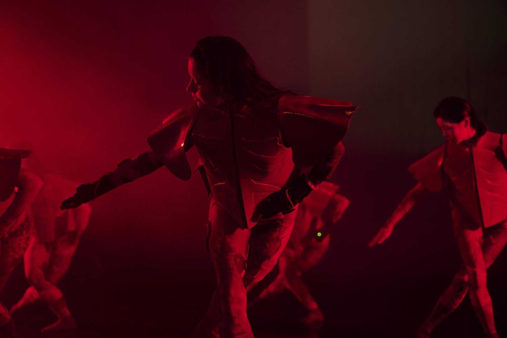 Mehrere Tänzerinnen bewegen sich in einer rot ausgeleuchteten Bühnenumgebung. Im Vordergrund steht eine Person in futuristisch wirkender, skulpturaler Kostümierung mit breiten Schulterelementen, die den Arm seitlich ausstreckt und den Blick nach unten richtet. Weitere Performerinnen sind unscharf im Hintergrund zu erkennen und greifen die Bewegung auf. Die Szene wirkt intensiv, technisch und leicht dystopisch.