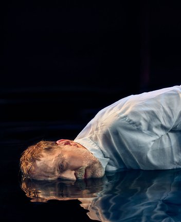 Nahaufnahme eines Mannes mittleren Alters vor schwarzem Hintergrund mit cineastischer Bildwirkung und warmer Lichtstimmung: Mit kurzem, zerzaustem braunem Haar liegt er auf einem wasserbedeckten Boden und blickt zum Betrachter. Er trägt ein weißes Hemd, sein Gesicht und Oberkörper spiegeln sich in der dunklen Wasserfläche. Sein Ausdruck wirkt erschöpft und nach innen gekehrt; die Szene vermittelt eine Atmosphäre von Rückblick und innerer Zerrissenheit. Sie evoziert die Figur des Hoffmann aus Jacques Offenbachs Oper Hoffmanns Erzählungen.