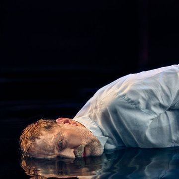 Close-up of a middle-aged man against a black background with a cinematic look and warm lighting: With short, tousled brown hair, he lies on a water-covered floor, looking toward the viewer. He wears a white shirt, and his face and upper body are reflected in the dark surface of the water. His expression appears exhausted and introspective; the scene conveys an atmosphere of reflection and inner turmoil. The scene evokes the figure of Hoffmann from Jacques Offenbach’s opera The Tales of Hoffmann.