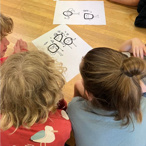 Mehrere Kinder sitzen im Kreis auf dem Boden und betrachten gemeinsam Zeichnungen von Tim Stadie. Auf den Blättern sind einfache Figuren zu sehen, die die „Operators“ der Oper Köln darstellen. Die Kinder diskutieren und entwickeln die Figuren weiter. Die Szene vermittelt konzentriertes, kreatives Arbeiten in einer Gruppe.