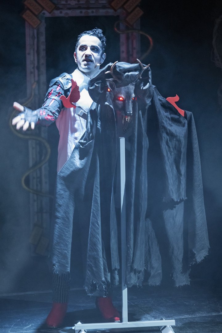 Auf einer düster beleuchteten Theaterbühne steht ein Darsteller als Kaspar aus der Oper „Freikugeln“ im Vordergrund. Er trägt ein kontrastreiches Kostüm aus weißen, schwarzen und roten Elementen mit auffälligen Details an Schultern und Ärmeln. Sein Gesicht ist stark geschminkt, mit blasser Haut und dunklen Augenpartien, was ihm ein unheimliches, fast dämonisches Aussehen verleiht. Mit ausgebreitetem Arm und intensiver Mimik wirkt es, als würde er eine beschwörende oder drohende Geste ausführen.
Neben ihm steht ein Objekt auf einem Gestell: eine schwarze, wolfsartige Figur mit Hörnern und leuchtend roten Augen, die aus einem dunklen Stoff hervorragt und eine bedrohliche Präsenz erzeugt. Der Stoff fällt wie ein Umhang nach unten und verstärkt die gespenstische Wirkung.
Die Bühne ist in kühles, bläuliches Licht und leichten Nebel getaucht, wodurch eine geheimnisvolle, unheilvolle Atmosphäre entsteht. Im Hintergrund sind angedeutete dekorative Elemente und geschwungene Formen zu erkennen, die den Eindruck eines magischen oder finsteren Ortes verstärken.