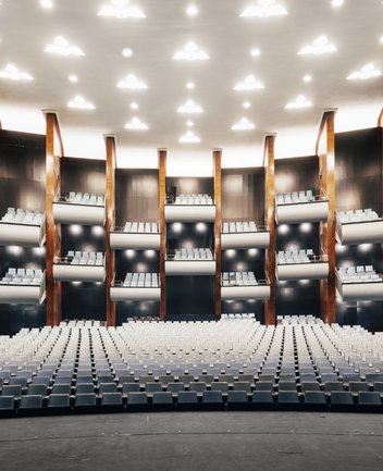Blick von der Bühne in den Zuschauerraum der Oper am Offenbachplatz mit ansteigenden Sitzreihen mit hellblauen Bezügen, mehreren geschwungenen Balkonen und warmen Holzvertäfelungen.