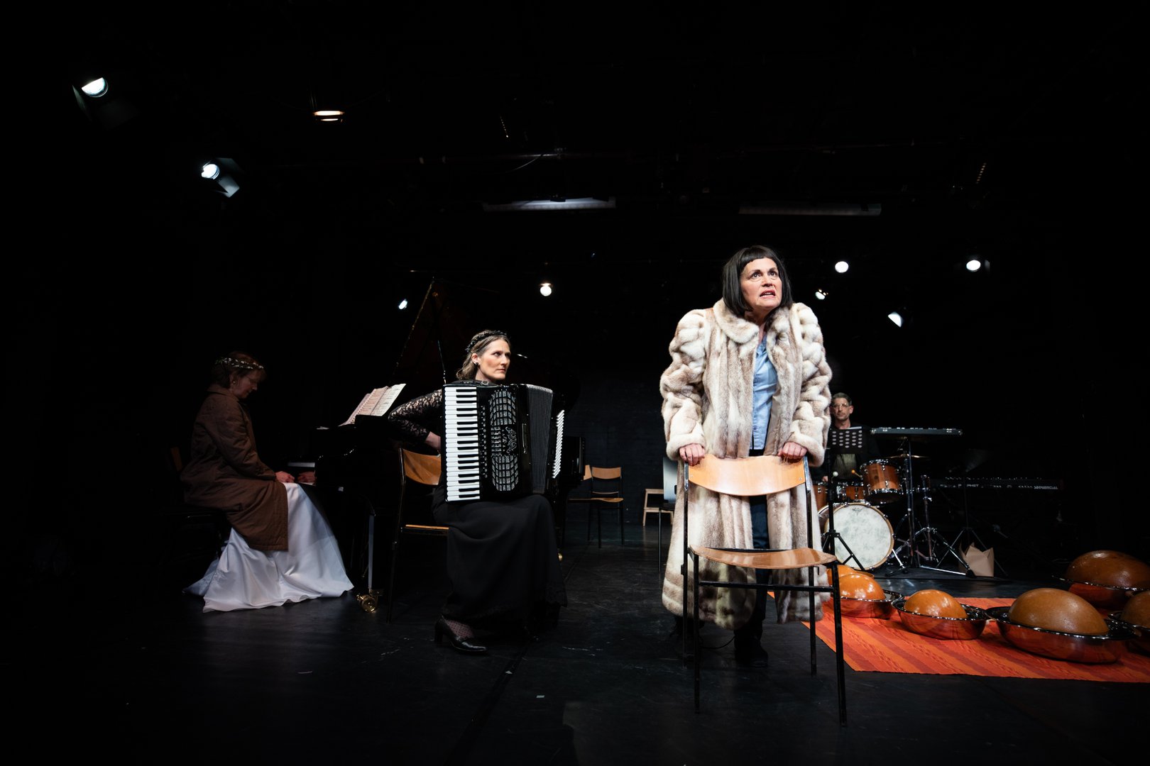 KS Dalia Schaechter in einem auffälligen Pelzmantel steht im Vordergrund einer Bühne und hält sich an der Lehne eines Stuhls fest, während sie intensiv nach vorne blickt. Links begleiten sie die Pianistin Theresia Renelt am Flügel und die Akkordeonspielerin Dorrit Bauerecker, rechts sitzt der Schlagzeuger Kurt Fuhrmann. Am Boden liegen mehrere halbkugelförmige Percussion-Instrumente auf einem Teppich. Die Szene ist dunkel beleuchtet und wirkt wie eine dramatische Kammermusik- oder Theateraufführung.