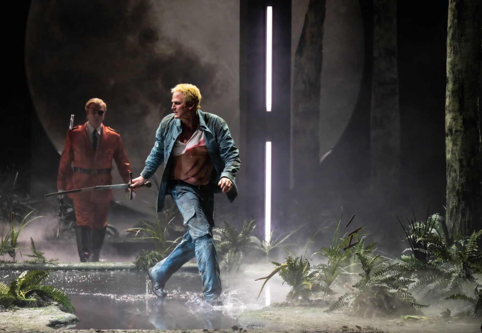 Eine Opernszene aus Wagners Die Walküre zeigt Wotan und Siegmund in einer nebelverhangenen, stilisierten Waldlandschaft. Im Vordergrund bewegt sich Siegmund energisch durch flaches Wasser, sodass Spritzer aufwirbeln. Er trägt eine offene Jacke und Jeans; sein T-Shirt ist zerrissen und gibt mehrere blutige Schnittwunden am Oberkörper frei. In der rechten Hand hält er ein Schwert. Seine Körperhaltung wirkt angespannt und zielgerichtet, als sei er auf der Flucht oder im Kampf.
Hinter ihm folgt Wotan in auffälligem, orangenem Mantel mit Gürtel und Augenklappe. Er geht aufrechter und kontrollierter, beinahe unbewegt im Vergleich zu Siegmund, was eine bedrohliche, überlegene Präsenz vermittelt.
Die Bühne ist von dichtem Nebel durchzogen, mit Farnen und niedrigen Pflanzen am Boden, die eine feuchte, naturhafte Umgebung andeuten. Vertikale Lichtstäbe im Hintergrund erinnern an stilisierte Baumstämme und erzeugen eine kühle, künstliche Waldästhetik. Dahinter ist ein großer, diffuser Mond sichtbar, der die Szene in ein gedämpftes, silbriges Licht taucht und die Spannung zwischen den Figuren verstärkt.