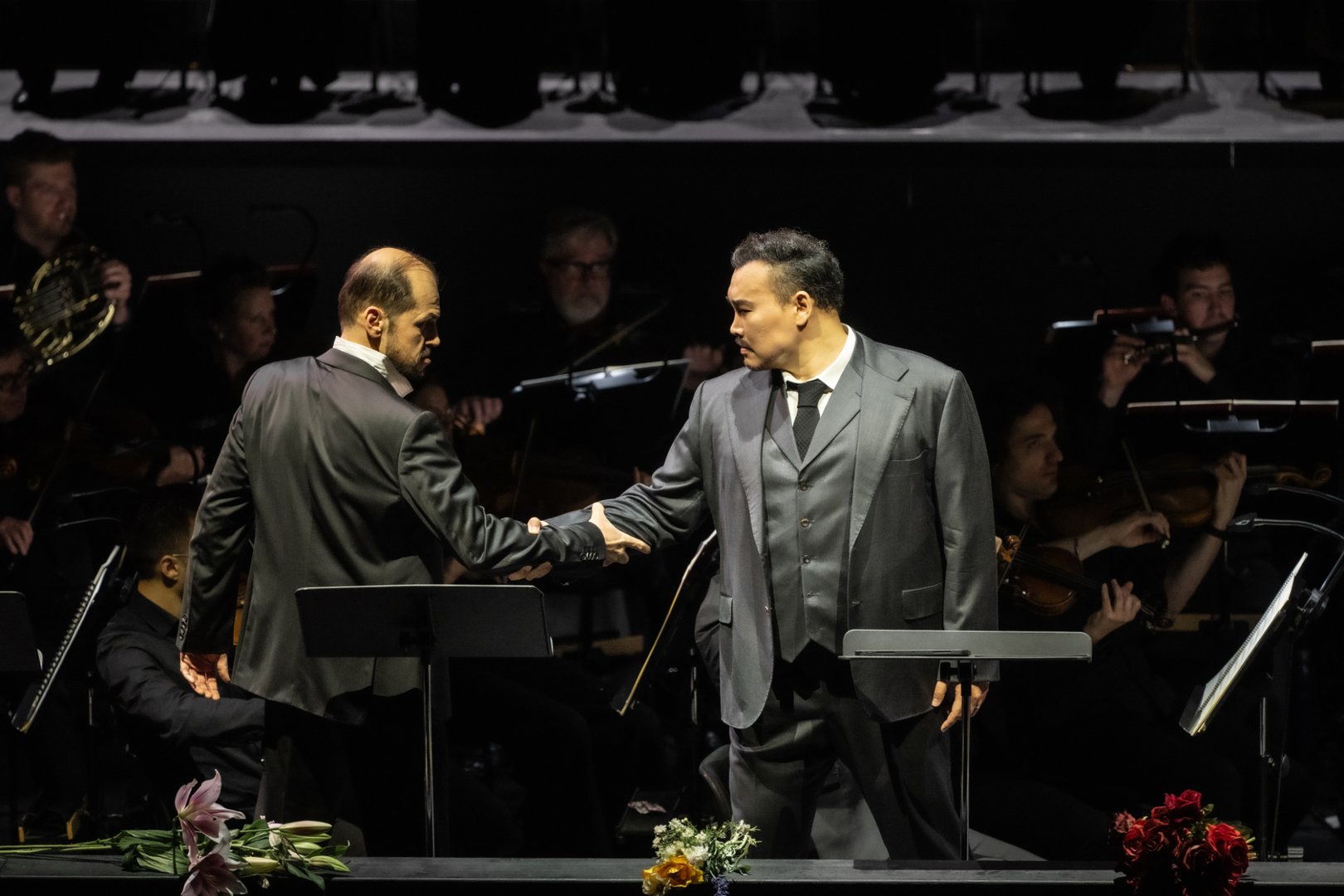 Auf der Bühne stehen Adrian Sâmpetrean (Don Ruy Gómez de Silva) und Young Woo Kim (Ernani) einander gegenüber und reichen sich die Hand. Beide tragen dunkle Anzüge und stehen an Notenpulten, während sie sich mit ernster, gespannter Mimik ansehen. Im Hintergrund ist das Gürzenich Orchester zu sehen, dessen Musiker*innen konzentriert ihre Instrumente spielen.