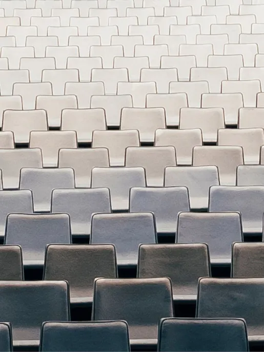 Blick auf zahlreiche, gleichmäßig angeordnete Sitzreihen im Zuschauerraum der Oper Köln am Offenbachplatz. Die hell gepolsterten Sitze bilden ein ruhiges, geometrisches Muster und vermitteln die Größe und Ordnung des Saals, während keine Personen anwesend sind.