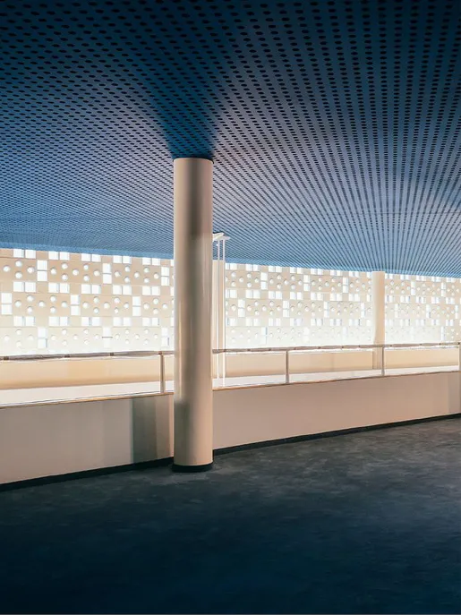 Zweites Geschoss des großzügigen Foyerbereichs der Oper Köln am Offenbachplatz mit hoher Decke und weiter Fensterfront. Die Wand ist von vielen kleinen runden und quadratischen Öffnungen durchzogen, durch die Tageslicht einfällt. Schlanke, helle Säulen tragen die Decke, die in einem kühlen Blauton gestaltet ist und einen Kontrast zur warmen Innenarchitektur bildet.