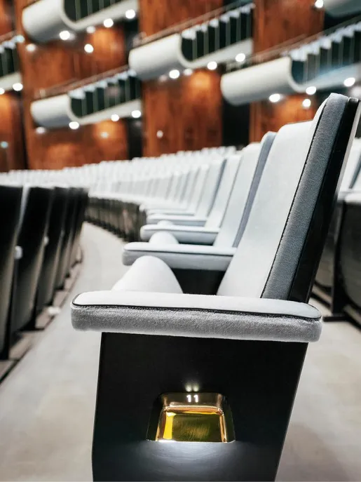 Seitliche Nahaufnahme einer Sitzreihe im Opernsaal der Oper Köln am Offenbachplatz mit gepolsterten Sitzen und integrierten Platznummern. Die Perspektive entlang der Reihe betont die Tiefe des Raums und die hochwertige Ausstattung.