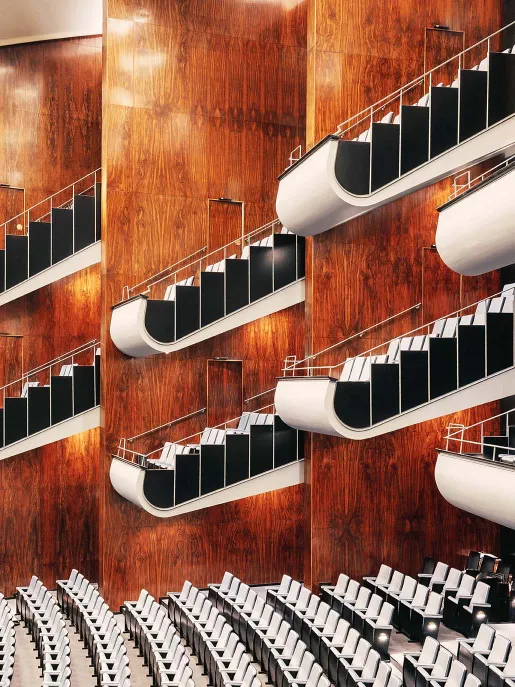 Several vertically stacked balconies of the Opera House auditorium at Oper Köln on Offenbachplatz. The curved balcony fronts, framed in white with black panels, stand out against the warm wood-panelled walls. Silver-coloured railings and the clean architectural lines create an elegant, contemporary atmosphere.