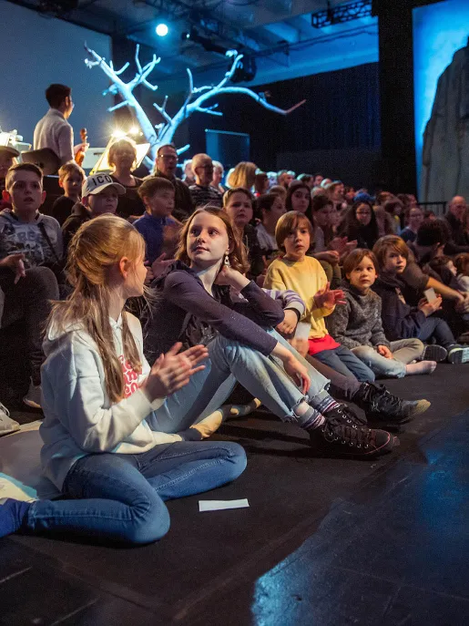 Several children are sitting on the floor directly in front of the stage, watching the performance attentively. Behind them, more children and adults are seated on tiered rows, following the performance. In the background, parts of the orchestra and a leafless stage tree are visible. The scene conveys the focused atmosphere of a family performance.