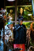 Maurizio di Mauro and Krista Krakel stand among withered plants in a whimsical magical workshop. On the left, Maurizio di Mauro, made up as a cat and wearing a hoodie and cap, looks mischievously towards the audience. On the right, Krista Krakel stands in a black, feather-like costume with a striking hairstyle and dark makeup. Behind them are a skeleton, a skull, plants, and other mysterious objects. Scene from The Satanarchaeolügenialcohellish Wishpunch.