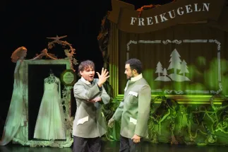 Scene from Freikugeln: Two performers in matching green-grey costumes with white details portray the character Max. The performer on the left uses German Sign Language, making Max accessible in two languages, while the other faces him attentively. Behind them, a stage set with the word “Freikugeln” and stylised fir trees fills the background. A white wedding dress stands in a decorative frame. Green lighting creates a fairy-tale-like, eerie atmosphere.