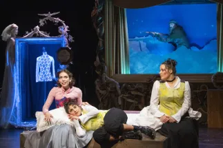 Scene from Freikugeln: In the foreground, Agathe (Ela Beysun) lies on a large wooden box with her head resting in Ännchen’s lap. She wears a yellow patterned top, a white blouse, and black trousers. She uses German Sign Language (DGS), making the character Agathe accessible in two languages. Ännchen sits on the left, speaking to both Agathes. On the right sits the singing Agathe. In the background, Head Forester Kuno walks through a snowy nighttime landscape carrying a rifle.
