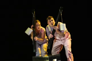 Scene from the children’s opera Max and Moritz: On a dark stage, Max and Moritz stand close together, looking startled towards the audience. Both wear light pink dungarees over pink T-shirts. Each holds a long black plank in front of them like a shield, with a white toilet paper roll hanging from a string attached to it. The scene suggests a moment of listening, hiding, or sneaking about under the spotlight.
