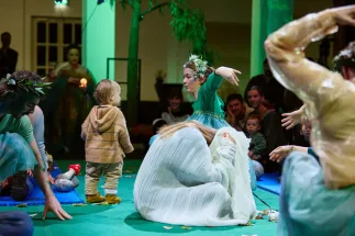 Scene from the opera for very young children Everything That Flies. In a circular performance space, a toddler stands among several performers wearing flowing costumes in shades of green, blue, and white. One performer, wearing a wreath of leaves, gestures expressively toward the child. Parents and children sit around the performance area, experiencing the action at close range. Natural elements, fabrics, and scattered leaves create a poetic and playful atmosphere.